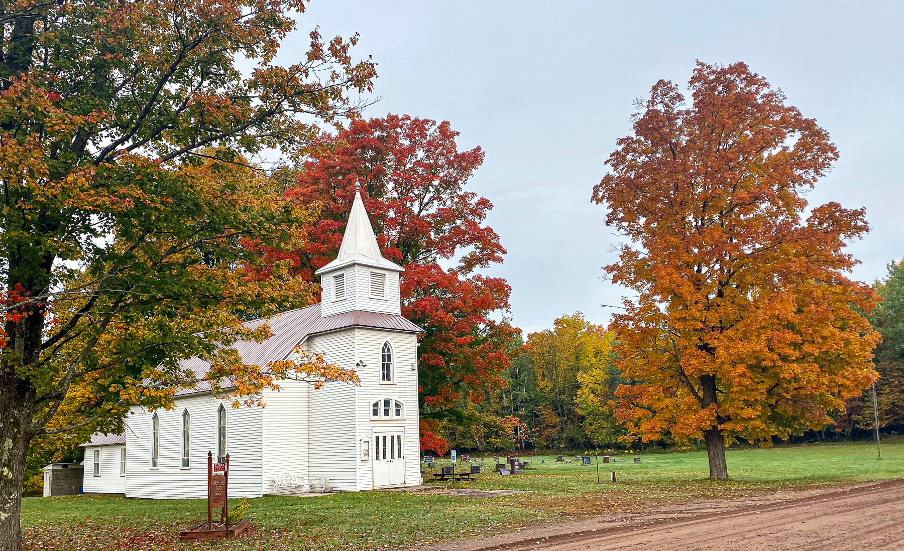 White church with steeple beside a grassy cemetery, framed by autumn red, orange, and yellow trees under a cloudy sky.